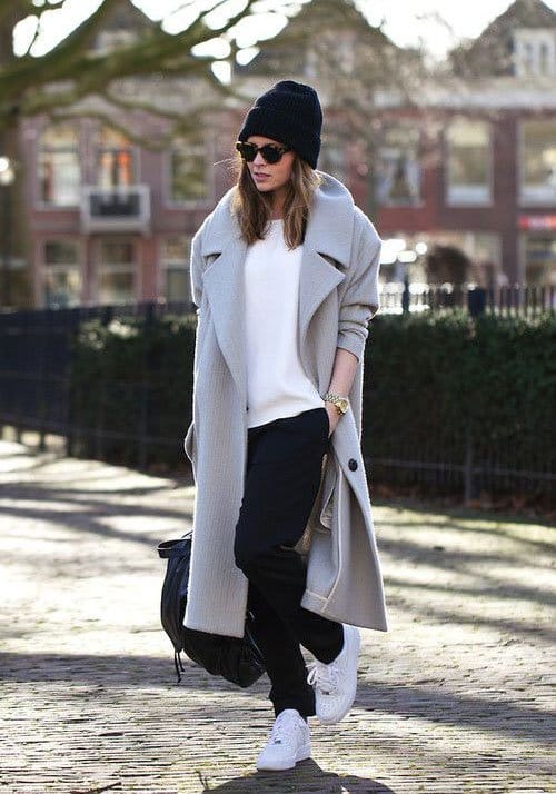 femme portant un jogging foncé, un haut blanc un manteau gris et des baskets blanches avec un bonnet noir et des lunettes de soleil