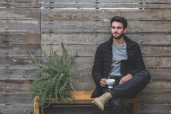 jeune homme assis avec une tasse à la main portant un jean foncé un t shirt gris des boots marron et une doudoune noire